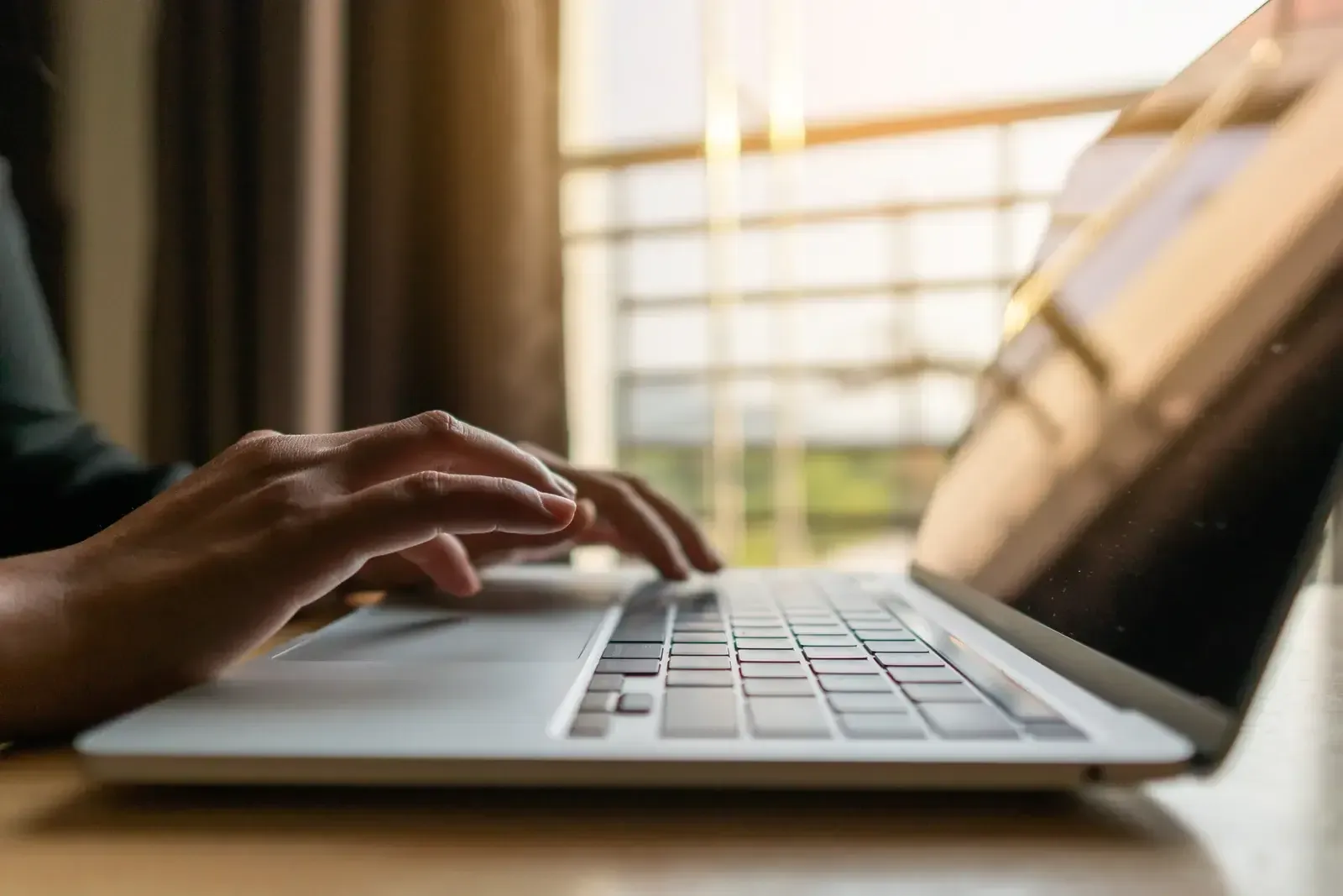 Une personne tape sur le clavier d’un ordinateur portable posé sur une table en bois, devant une grande fenêtre baignée de lumière chaude